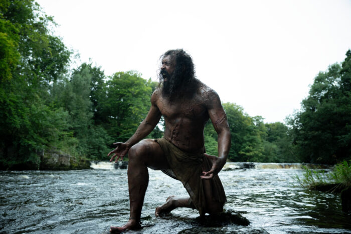 A scarred man emerges from a shallow river in a tense outdoor scene from “28 Years Later: The Bone Temple.”
