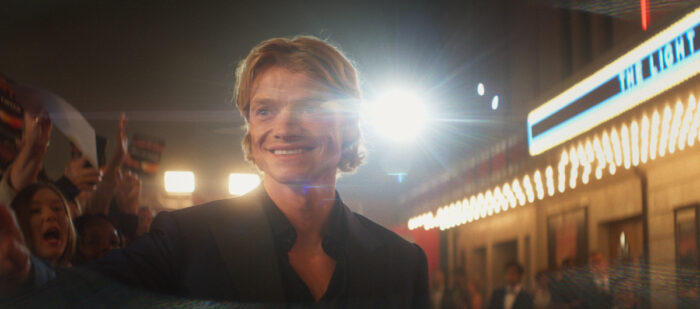 Tom Rhys Harries smiles on the red carpet surrounded by flashing cameras and fans in a glamorous Hollywood scene from “Clayface,” before his character’s dark descent.
